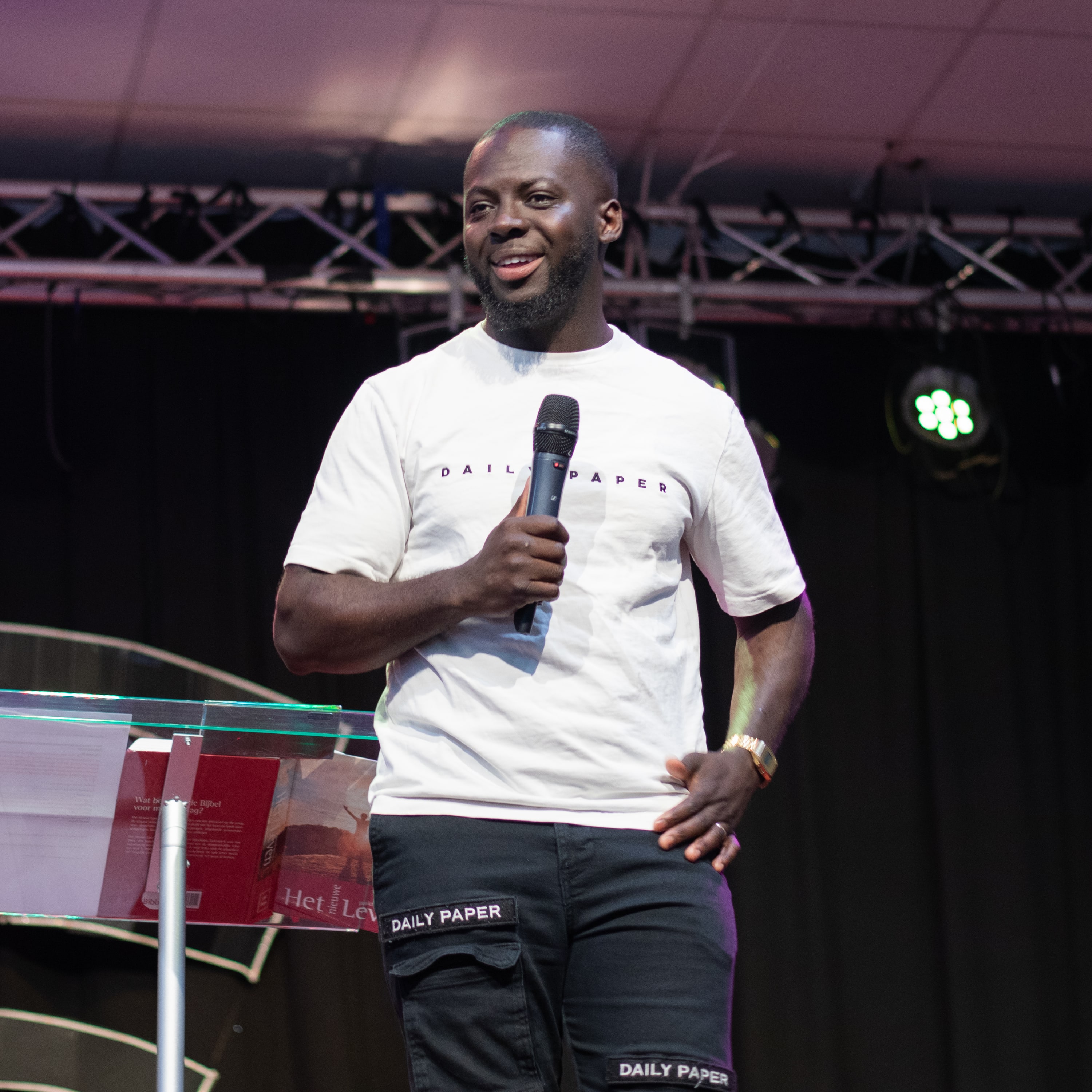 Godwin Arhin staat lachend met een microfoon in de hand op een podium, in wit T-shirt naast een doorzichtige lessenaar met een opengeslagen boek