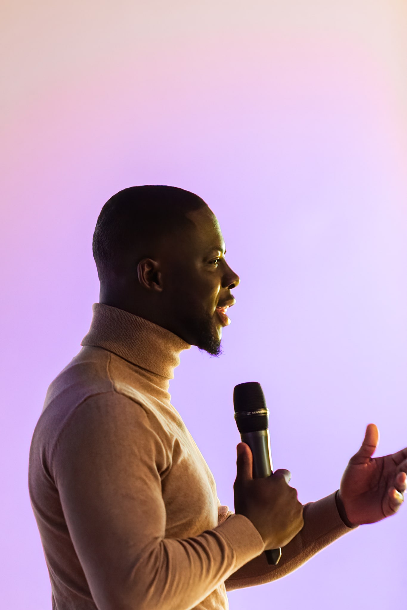 Godwin Arhin spreekt in profiel met een microfoon in zijn hand, in beige coltrui, gebarend tegen een paarsroze achtergrond